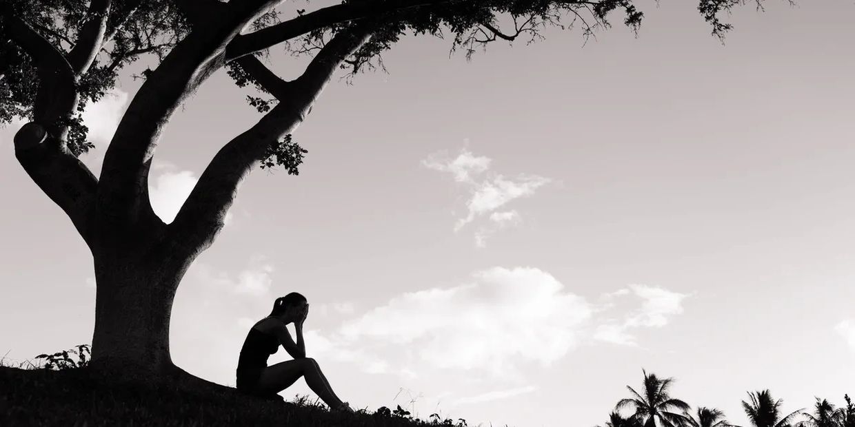 A person sits alone under a large tree, appearing contemplative or distressed.