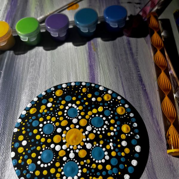 Colorful dot mandala painting on black surface with paint pots and tools.