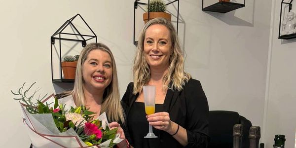 Two women smiling, one holding flowers, the other a drink, in a decorated room.