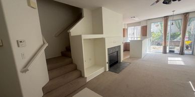 Empty living room with staircase and fireplace leading to a bright kitchen area.