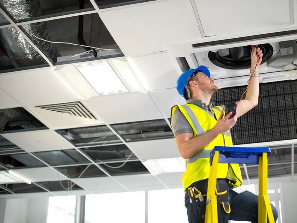 A worker in a hard hat and safety vest inspects a ceiling air conditioning unit