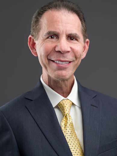 Smiling middle-aged man in a suit with a yellow tie against a gray background.