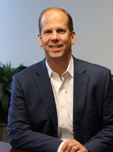 Smiling middle-aged man in a navy suit and white shirt seated indoors.