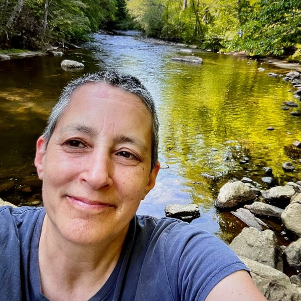 Person smiling near a calm river in a forest.
