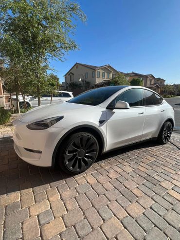 White Tesla Model Y parked on a brick driveway in a suburban neighborhood.