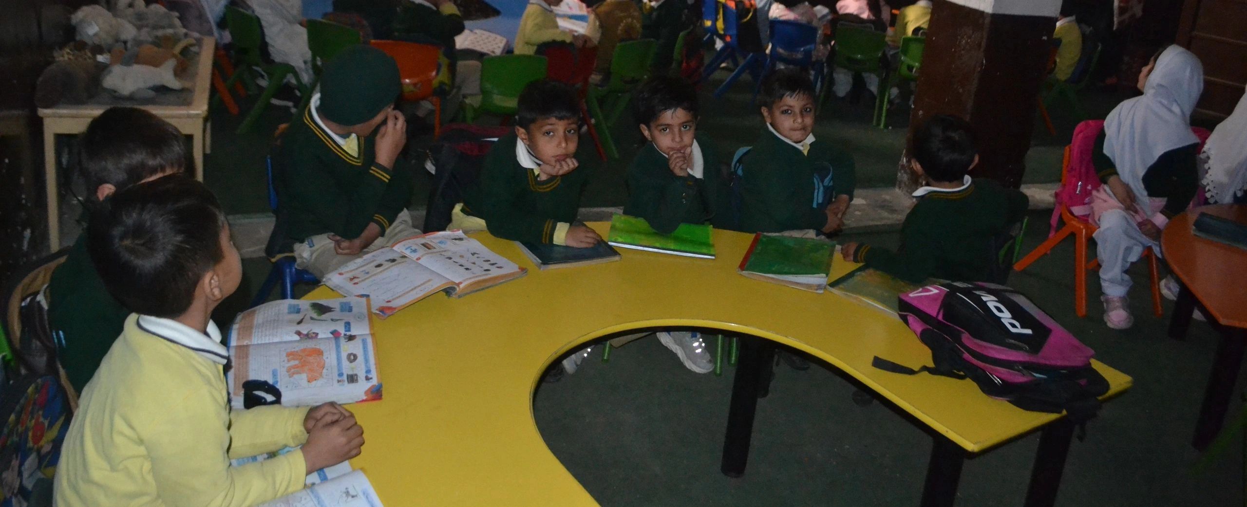 Children in school uniforms sitting around a yellow table with books open.