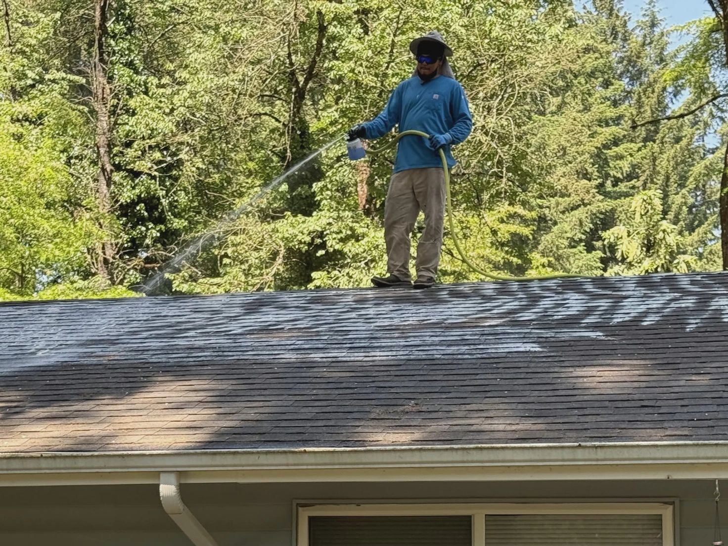 Worker spraying cleaning solution across a shingle roof.