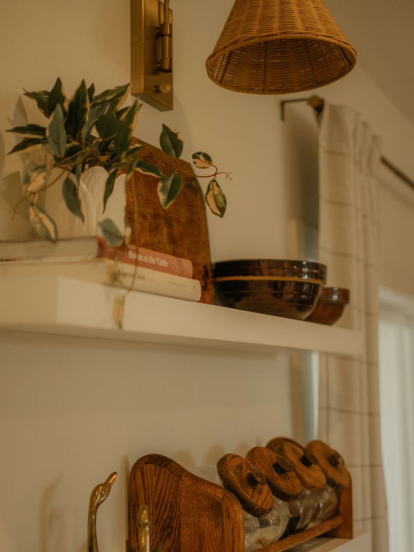 Cozy kitchen shelves with plants, books, wooden jars, and brass decor under warm lighting.