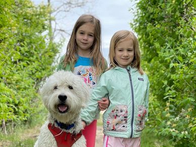 Two girls stand outdoors with a fluffy white dog on a green path.