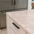 "Close-up of a modern kitchen island with a marble countertop, sleek black handle on a white cabinet drawer, and a stainless steel refrigerator in the background