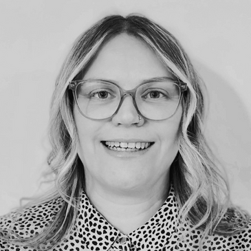 Black and white image a woman with wavy hair, wearing glasses, smiling friendly.
