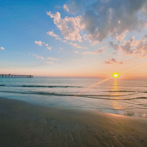 Sun setting over calm beach waves, creating soft pink and blue tones. Pier visible in the distance.
