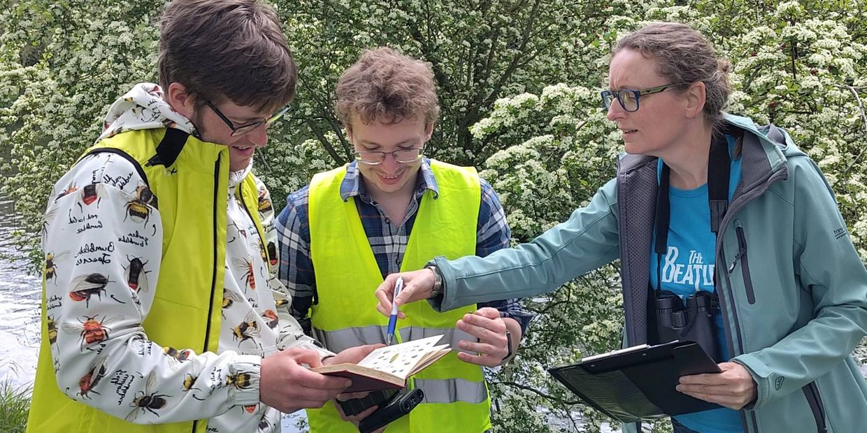 Three people outdoors examining a book and notes, wearing bright vests and casual jackets.