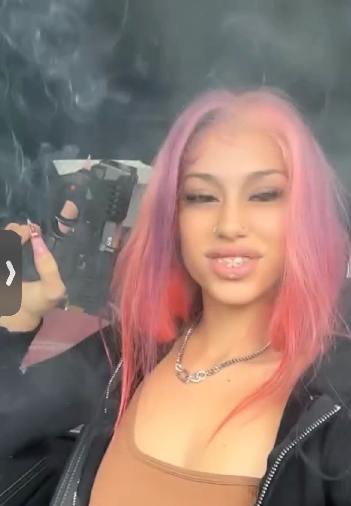 Woman with pink hair and piercings smoking a cigarette indoors.