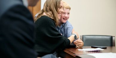 A woman hugs a smiling boy at a table in an office setting.