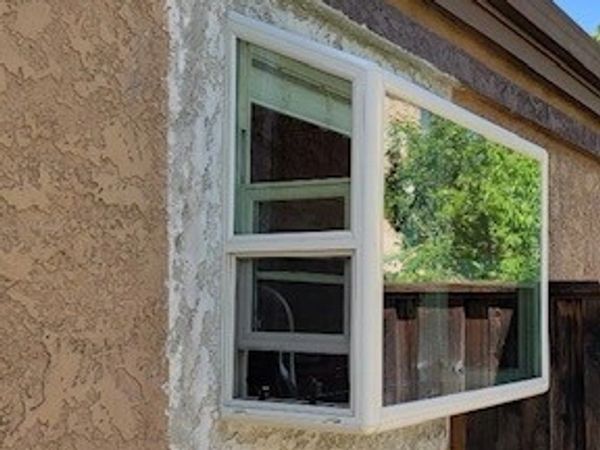 A white-framed bay window on a stucco wall reflecting greenery and a wooden fence.