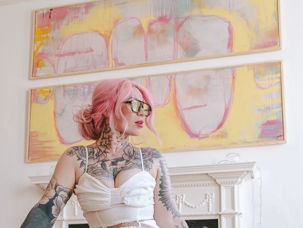 Tattooed woman with pink hair and white outfit sitting on a green ottoman.