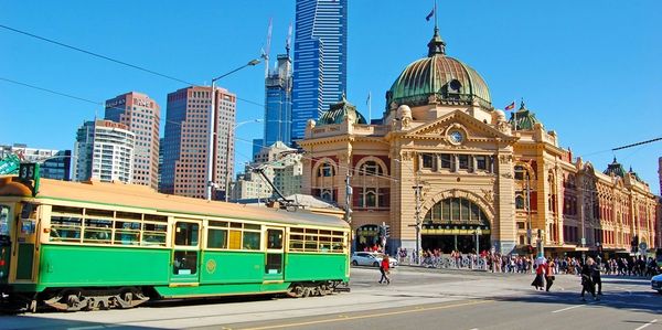 A green tram passes Flinders Street Station in Melbourne with city skyscrapers behind.