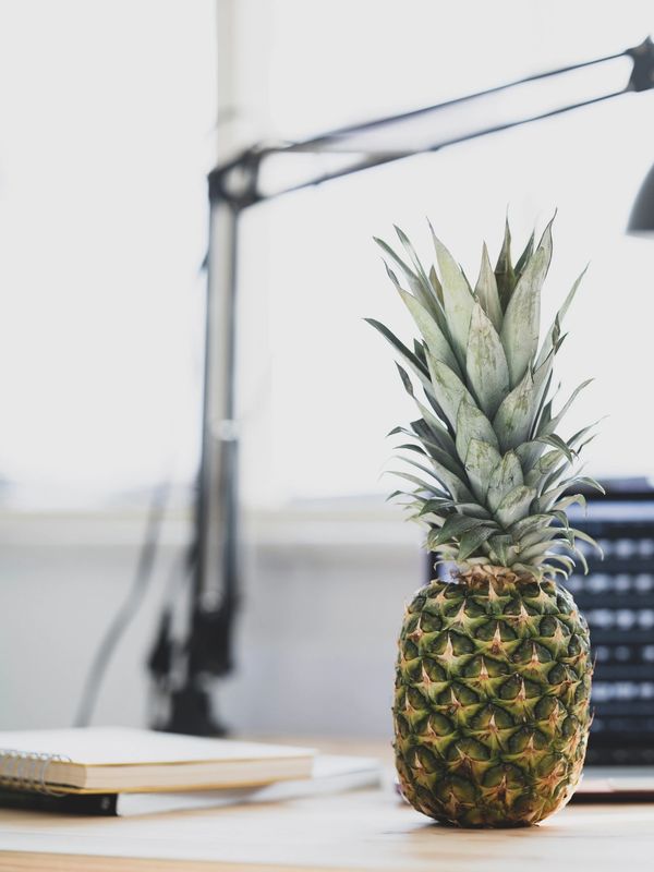A pineapple sits on a wooden desk with a notebook and lamp in the background.