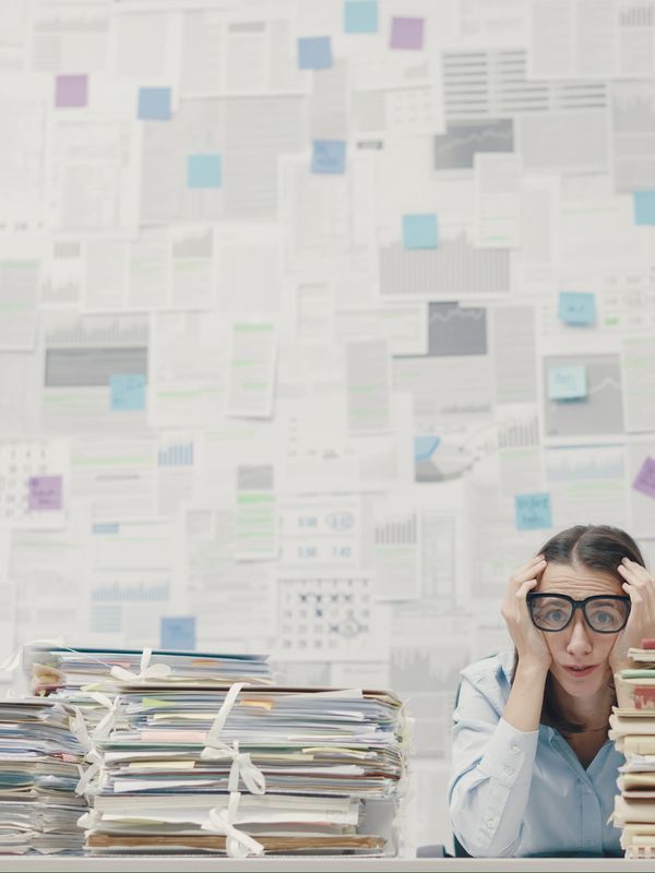 Overwhelmed woman surrounded by stacks of paperwork and notes on the wall.