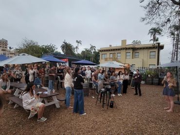 Outdoor gathering with people listening to a speaker near picnic tables and umbrellas.