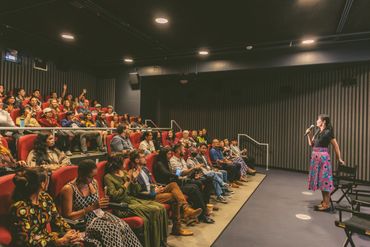 A woman speaking to an attentive audience in a theater setting.