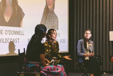 Three women engaged in a panel discussion on stage with a projected presentation behind them.
