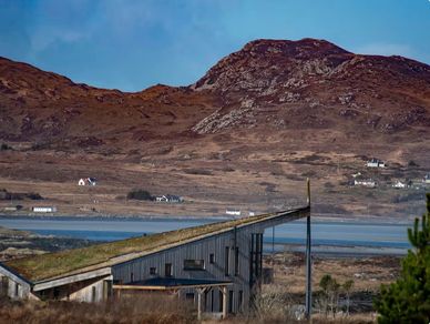 Rustic house with a green roof by a coastal landscape and hills.
