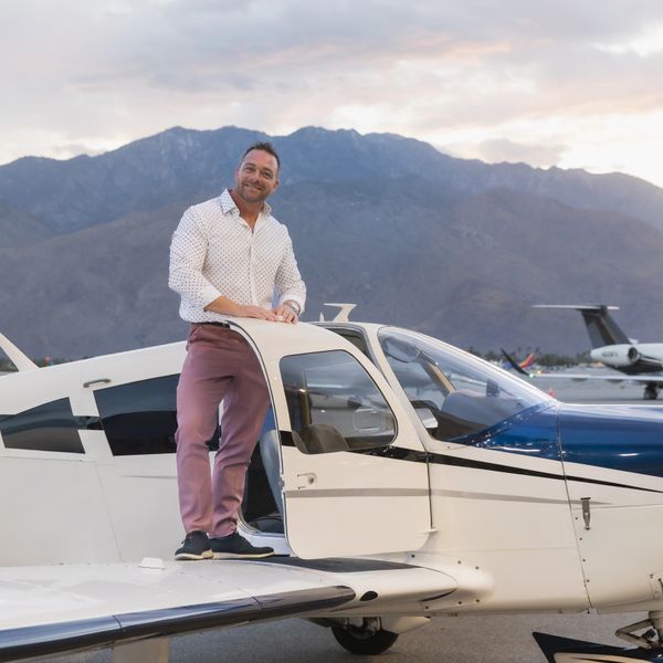 Man standing on the wing of a small airplane smiling with mountains in the background.