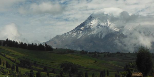 Snow-capped mountain towering over lush green fields under a cloudy sky.