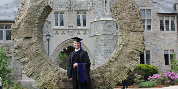 Graduate in cap and gown poses inside a large circular stone sculpture on campus.