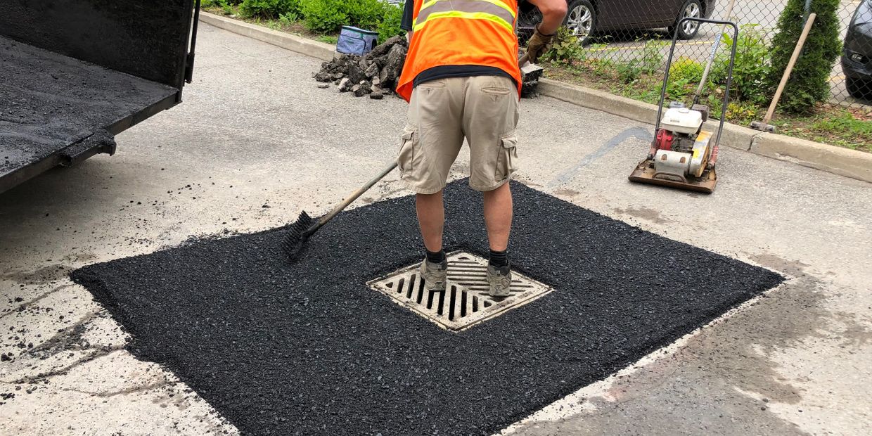 Worker in an orange vest paving asphalt around a drain on a street.