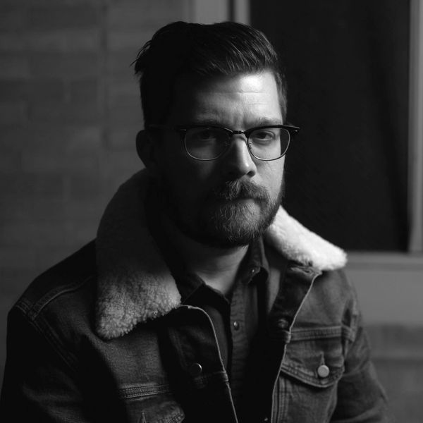 Black and white portrait of a man with glasses and a beard wearing a shearling jacket.