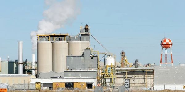 Industrial factory with multiple silos and a water tower emitting smoke.