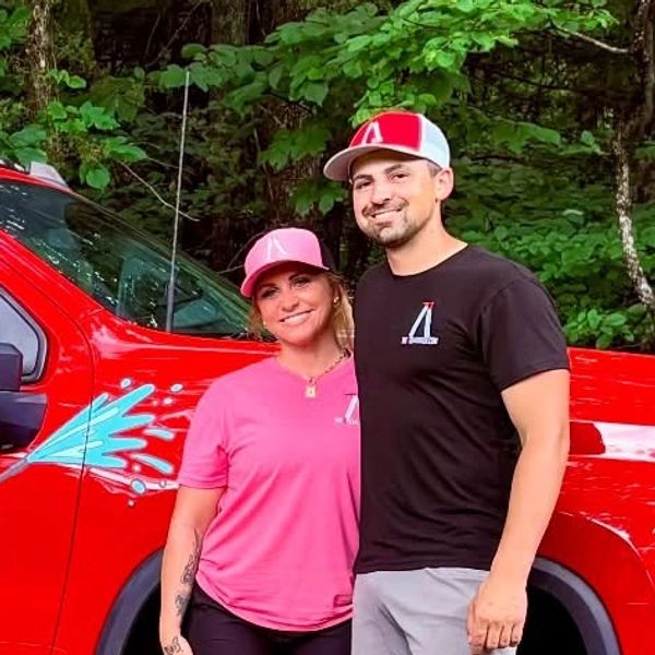 A smiling couple stands next to a red truck in a wooded area.