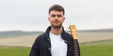 A man standing in a green field holding an acoustic guitar.