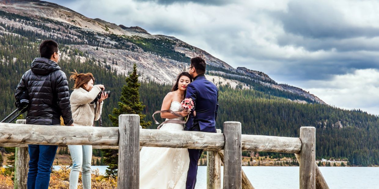 A couple poses for wedding photos by a lake with mountains in the background.