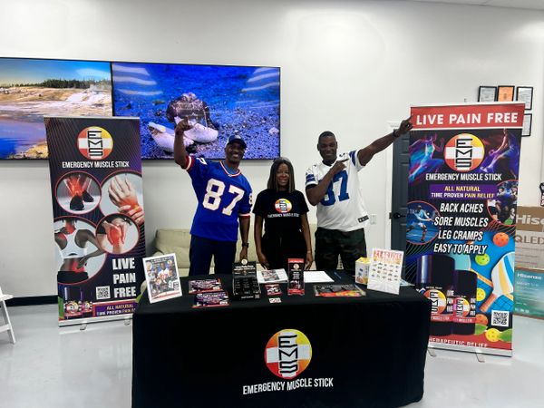 Three people promoting Emergency Muscle Stick at an event booth with banners and products.