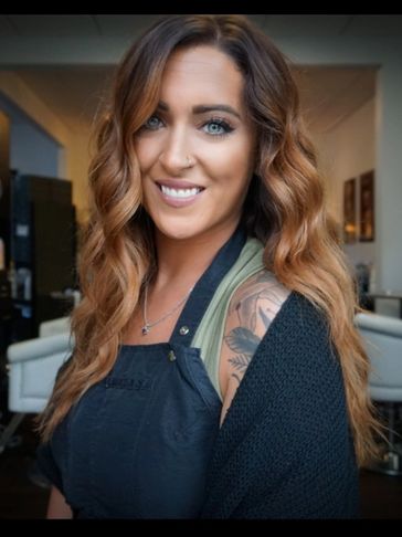 Smiling woman with wavy hair and a visible tattoo, wearing a black apron and cozy sweater.