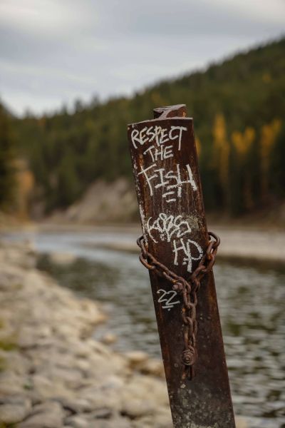 Rusty pole by a river with graffiti saying 'Respect the Fish' and a chain wrapped around it.