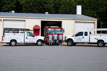 Four men standing between two white work trucks outside a building.