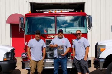 Three men stand in front of a fire truck, one holding a plaque.
