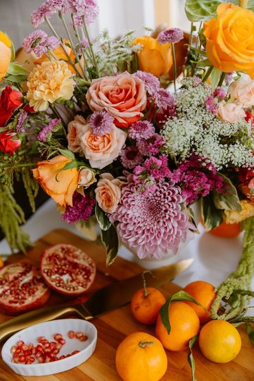 A vibrant flower bouquet with roses and chrysanthemums beside fresh oranges and pomegranate seeds.