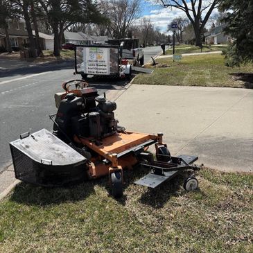 A lawn mower parked on the grass near a sidewalk and a street.
