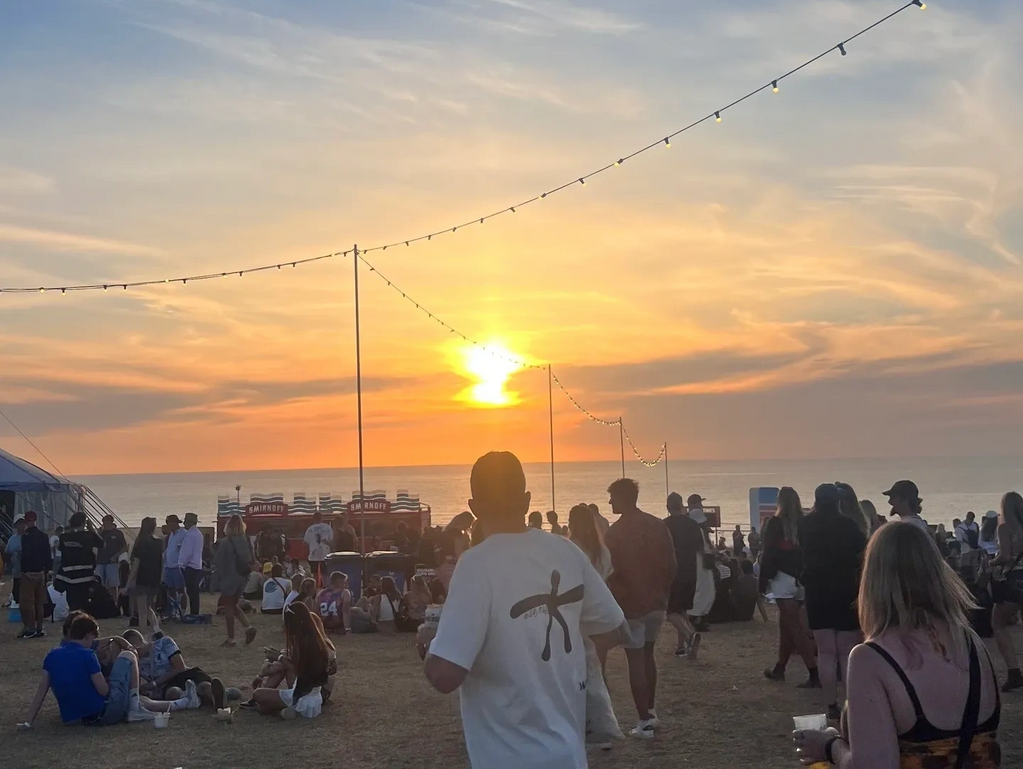 People enjoying a sunset beach festival with string lights overhead.