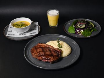 A steak dinner with mashed potatoes, salad, soup, and a glass of juice.