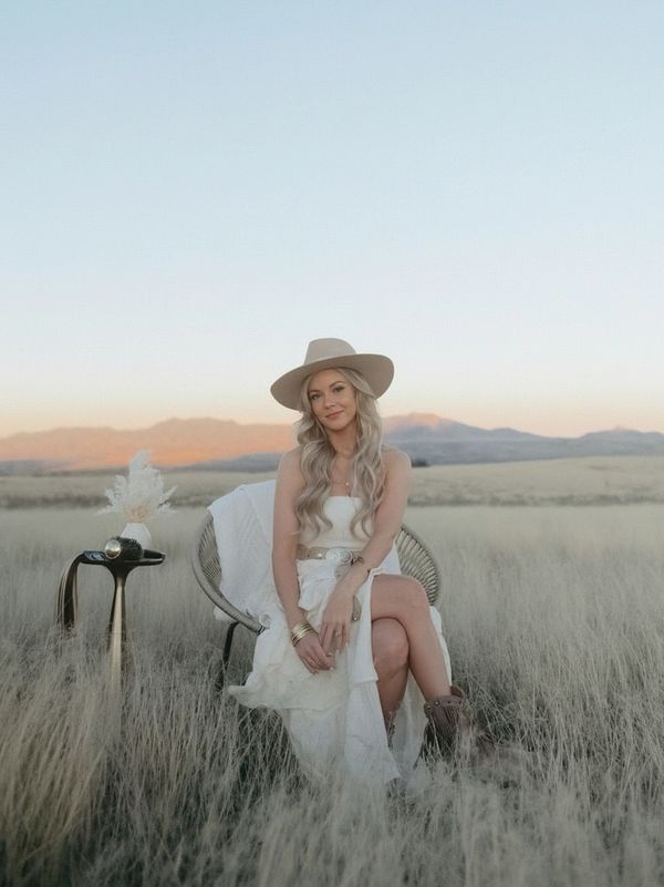 Woman in white dress and hat sitting in a field at sunset.