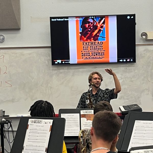 A man teaches music to a classroom, holding a saxophone and gesturing.