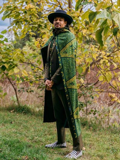 Man wearing a green patterned cape and a black hat in a forest.