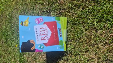 Children's book titled 'Mamie &amp; Rob and the Red Envelope' displayed outdoors on grass.